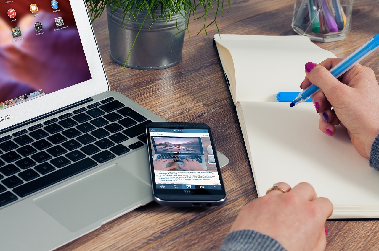 A person writes in a notebook with a blue pen at a desk, next to an open laptop, a smartphone, and colored pencils—an organized setup perfect for a REAL ESTATE VIRTUAL ASSISTANT or top sales representative assistant.