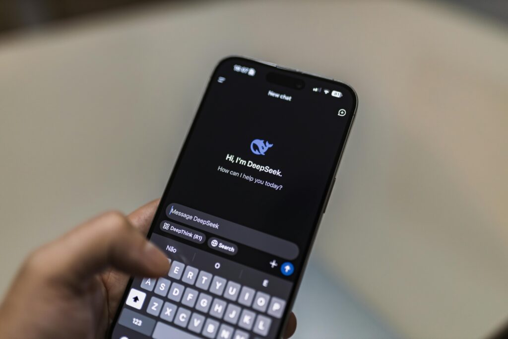 A person holds a smartphone displaying a chat with DeepSeek AI, typing a message to their REAL ESTATE VIRTUAL ASSISTANT on the devices keyboard.