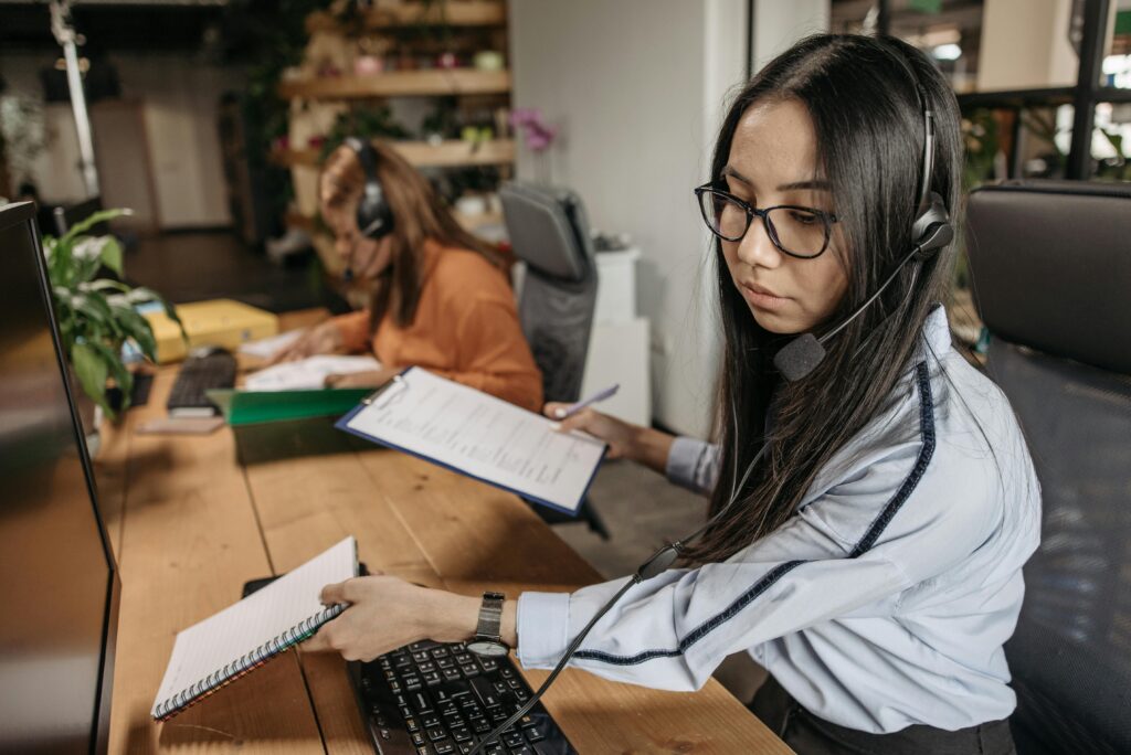Two women, including a SALES REPRESENTATIVE ASSISTANT, work at desks. The woman in the foreground reviews documents and types, while the other writes in a notebook. Both are focused on their tasks in an office setting.