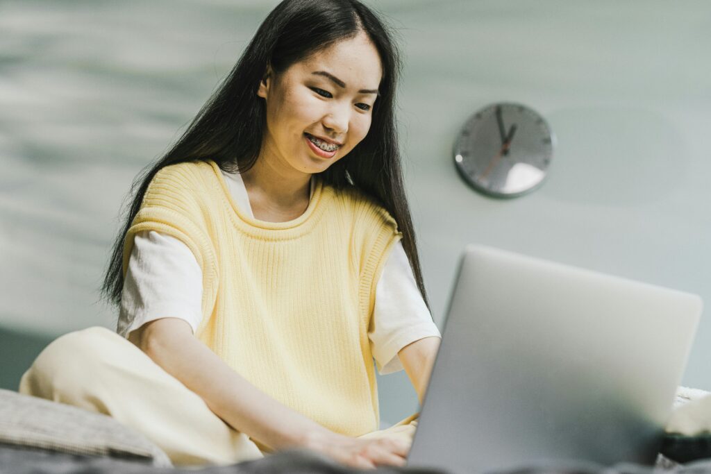 A young woman with long dark hair and braces, wearing a yellow sweater, sits on a bed and smiles while using her laptop—perhaps exploring virtual assistant services. A round wall clock is visible in the background.