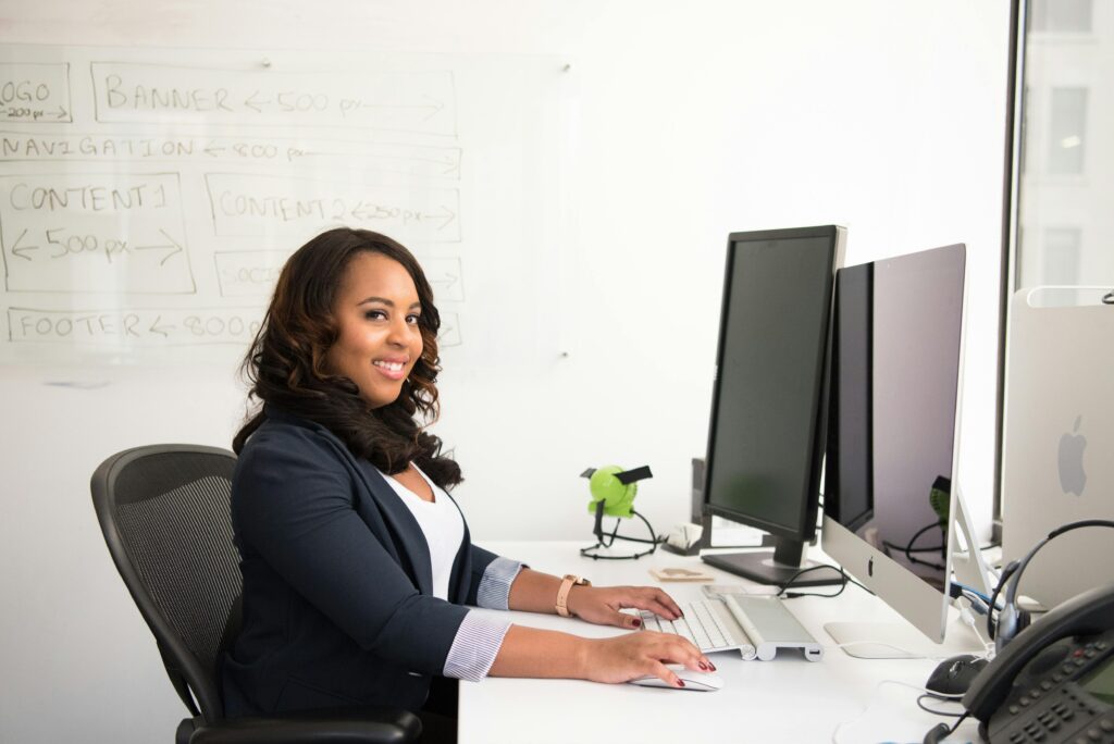 A woman in business attire sits at a desk, smiling at the camera while typing on a keyboard, with two large monitors and a whiteboard with notes in the background.